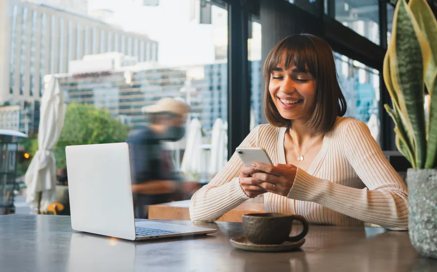 A smiling young woman sitting at a table in an café with a coffee cup and browsing her smartphone and laptop.